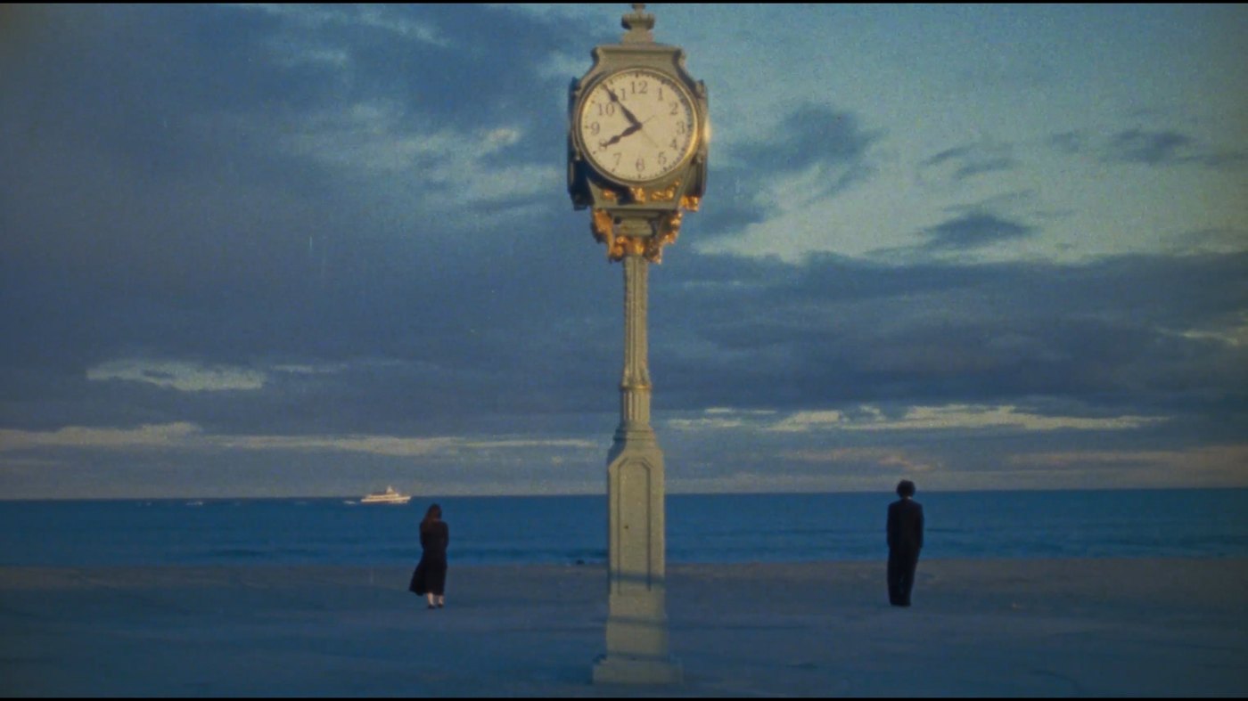 A clock tower on an empty beach at dusk, two figures standing apart from one another