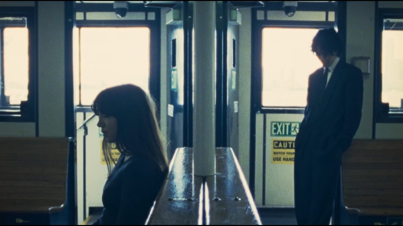 A woman and a man stand at opposite ends of a ferry cabin, backs to one another, harbour light behind them