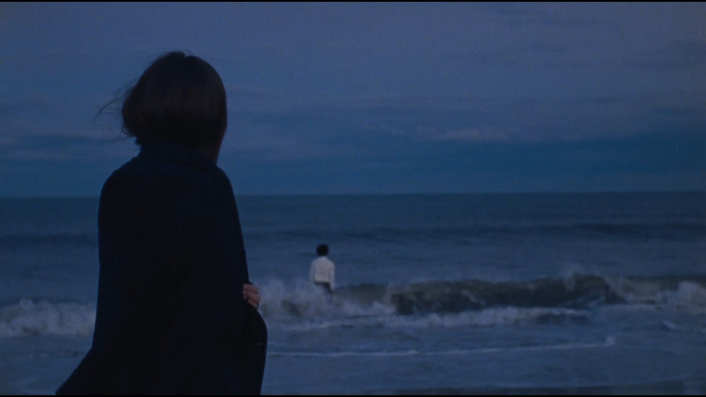 A woman watches from the shore as a man walks into the sea at blue hour